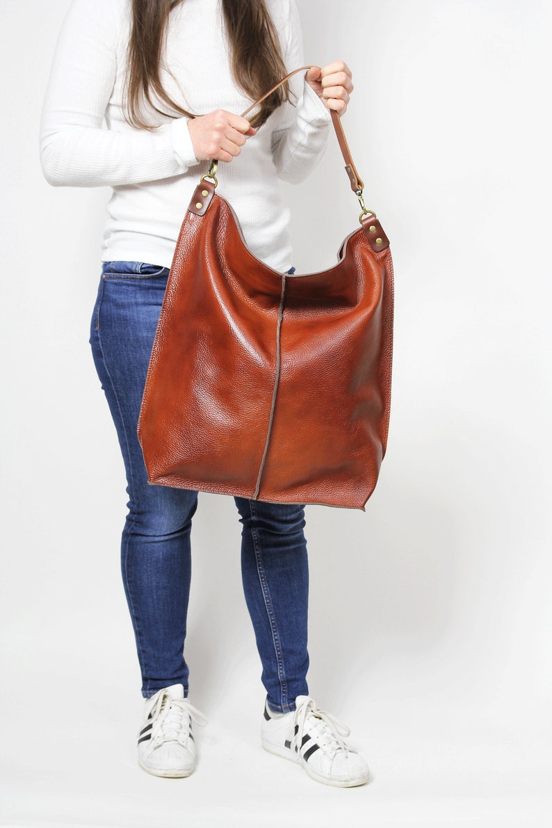 Peut inclure: Un grand sac fourre-tout en cuir marron avec une longue bandouli&egrave;re. Le sac est port&eacute; par une personne v&ecirc;tue d'un jean bleu et de baskets blanches.