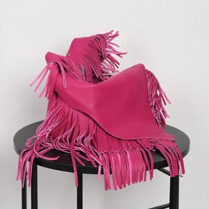May include: A vibrant hot pink handbag featuring a textured surface and long, flowing fringe detailing that hangs from the edges. The bag has a structured yet draped appearance with a single visible metallic stud accent near the top handle area.