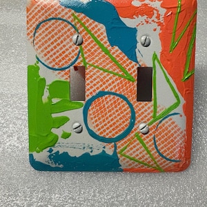 May include: A square, hand-painted light switch cover with a vibrant abstract design. The cover features a white base with orange, teal, and green paint. Geometric shapes and patterns, including circles and triangles, are incorporated into the design. Two toggle switch openings are visible.