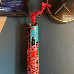May include: A cylindrical wall vase with a vibrant, hand-painted design. The vase features a black base, turquoise accents, and a red and pink pattern. Two red calla lilies are placed inside the vase. A gold-rimmed clock is visible in the background.
