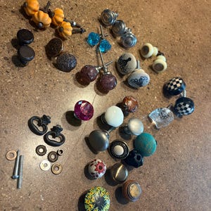 May include: A collection of decorative cabinet knobs in various styles and colors. Includes knobs with pumpkin, floral, and geometric designs, as well as knobs with clear, blue, and black accents. Hardware and screws are also included.