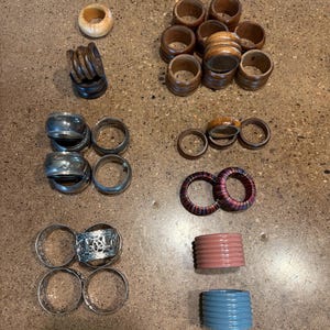 May include: A collection of napkin rings in various styles and materials. Includes wooden, silver, and colorful plastic rings. Some are plain, while others have decorative details or patterns. A variety of colors and textures are visible.