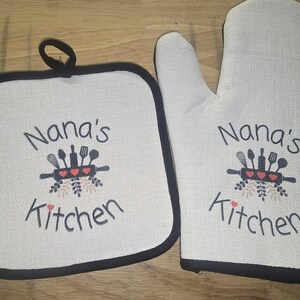 May include: A cream-coloured pot holder and oven mitt set. Both items have a black border and feature the text "Nana's Kitchen" with a graphic of kitchen utensils and hearts.