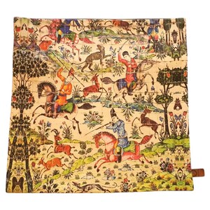 May include: Square cushion featuring a detailed, colourful illustration of a hunting scene. The artwork depicts figures on horseback, deer, dogs, and trees, set against a beige background. A brown leather tab is attached to the side.