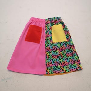 May include: A vibrant skirt with an elasticated waistband. One half is bright pink with a red pocket, the other features a rainbow leopard print with a yellow pocket. A playful and colourful garment.