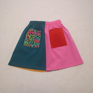 May include: A vibrant skirt featuring a split design. One half is teal with a rainbow leopard print pocket, the other is pink with a red pocket. It has an elasticated waistband and a yellow hem.