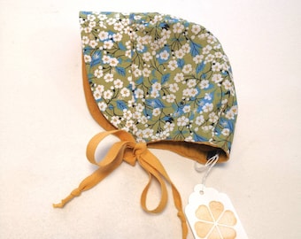 May include: A floral print baby bonnet with a mustard yellow tie. The bonnet features a pattern of white flowers and blue leaves on a green background. The interior and tie are mustard yellow. A tag with the text "Y-KAIROS" is attached.