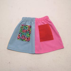 May include: A two-tone skirt in light blue and pink. The light blue section features a pocket with a colourful leopard print, and the pink side has a red pocket. The skirt has an elasticated waistband.