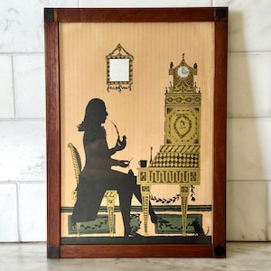 May include: A framed silhouette art piece depicting a person smoking a pipe at a desk with a clock and mirror. The artwork features a dark silhouette against a tan background, framed in dark wood. The desk and clock have gold accents.