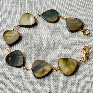 May include: A handmade bracelet with heart-shaped, polished stones in brown, tan, and blue-grey. The stones are linked by delicate gold-coloured wire and secured with a gold-tone clasp. The bracelet is displayed on a neutral-coloured fabric.