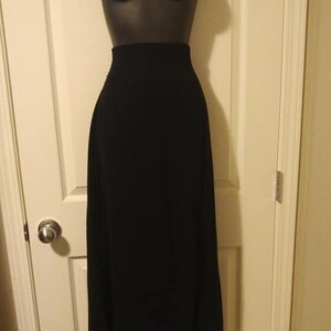 May include: A long, black skirt displayed on a mannequin. The skirt is floor-length and has a high waist. The fabric appears to be a solid, opaque material. The mannequin is a dark color, and the skirt is hanging in front of a white door.