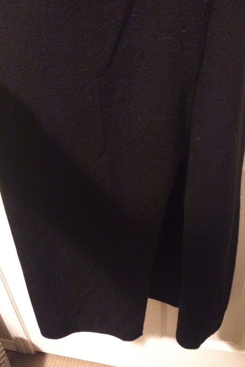 May include: Close-up of a black garment, possibly a skirt or dress, with a side slit. The fabric appears to be a soft, knit material. The hemline is slightly curved. The garment is hanging against a white door frame.