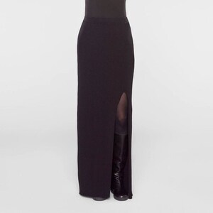 May include: A black, floor-length skirt with a high slit on the left side. The skirt is ribbed and form-fitting, with a simple, elegant design. The model is wearing black boots.