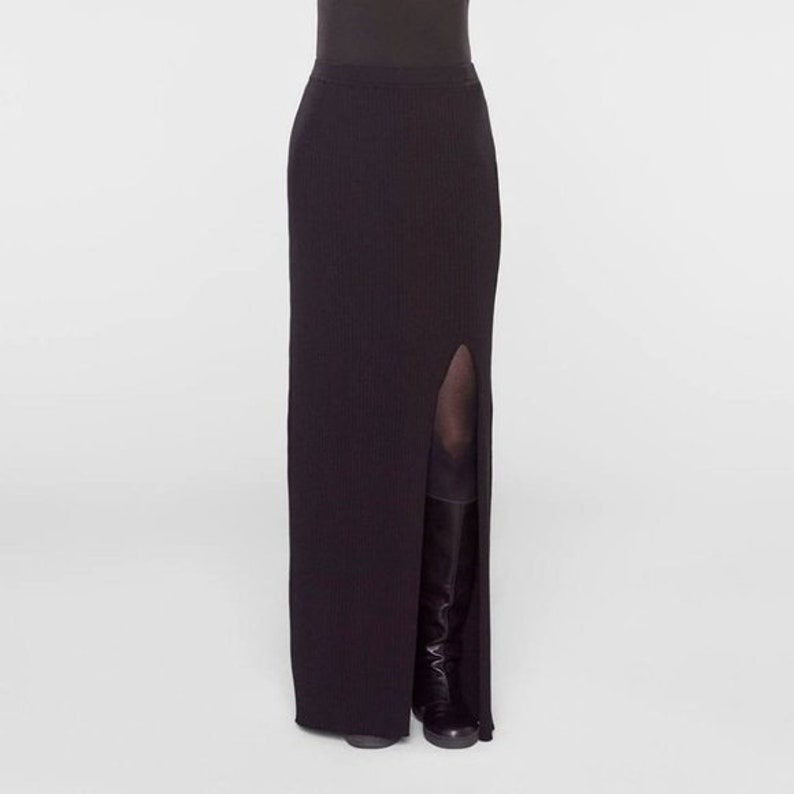 May include: A black, floor-length skirt with a high slit on the left side. The skirt is ribbed and form-fitting, with a simple, elegant design. The model is wearing black boots.