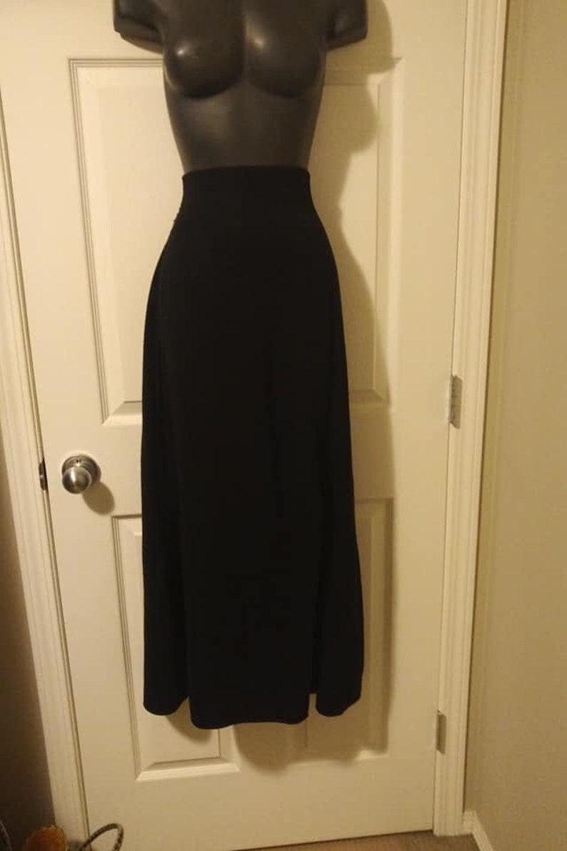 May include: A long, black skirt displayed on a mannequin. The skirt is floor-length and has a high waist. The fabric appears to be a solid, opaque material. The mannequin is a dark color, and the skirt is hanging in front of a white door.