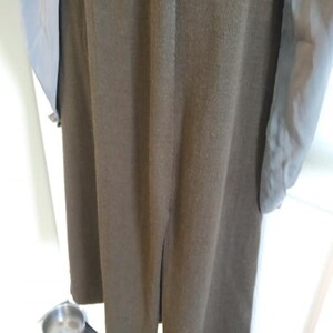 May include: A long, olive green coat with a split at the hem. The coat has long sleeves with a light blue lining. The coat is hanging on a wooden hanger. A small, silver bowl sits on the floor.