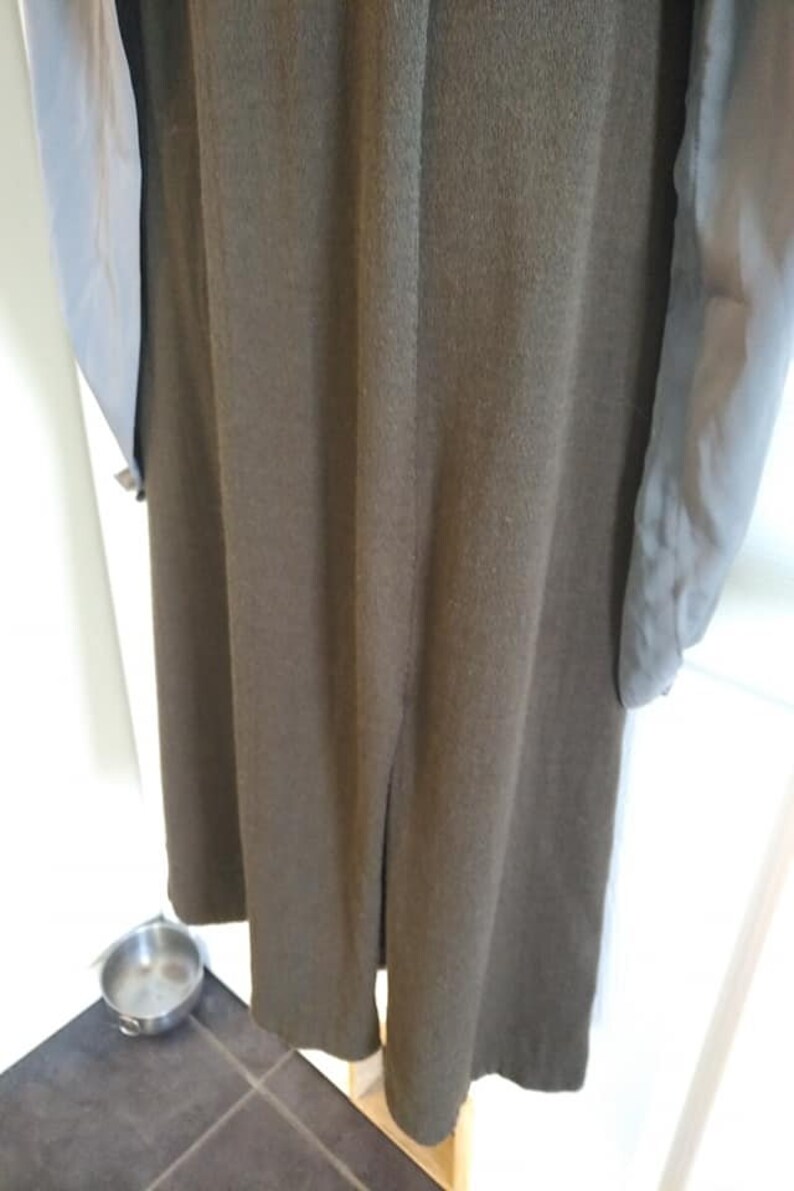 May include: A long, olive green coat with a split at the hem. The coat has long sleeves with a light blue lining. The coat is hanging on a wooden hanger. A small, silver bowl sits on the floor.