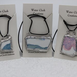 Può includere: Tre collane con cornici argentate che contengono pezzi di vetro di diversi colori. I pezzi di vetro sono di forme e dimensioni diverse. Le collane sono tutte su cordini neri e sono appese a carte bianche con il testo "Water Clock Creations" su di esse.