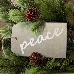 May include: A rustic metal tag with the word "peace" written in white cursive lettering. The tag is surrounded by green pine branches and brown pine cones.
