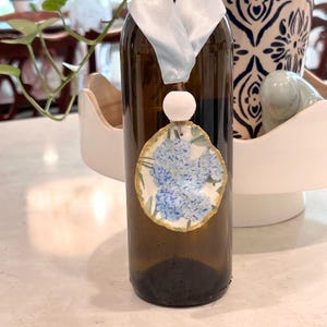 May include: A brown glass bottle adorned with a light blue ribbon and a decorative pendant. The pendant features a floral design with blue hydrangea blooms and a gold-painted edge. A white bead hangs between the ribbon and pendant.