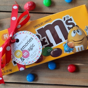 May include: A small box of M&M's chocolate candies wrapped in a red ribbon with white polka dots and a tag that says "Let's make some memories & magic on the field today".