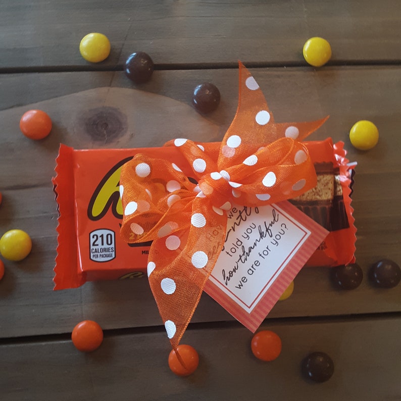 May include: A chocolate candy bar wrapped in orange and brown paper with a white and orange polka dot ribbon tied around it. A small card with the text "Have we told you how thankful we are for you?" is attached to the ribbon.