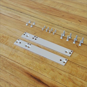 May include: Two silver metal mounting plates with multiple holes, along with an assortment of silver screws, are arranged on a wooden surface. The plates are rectangular and have a series of evenly spaced holes.
