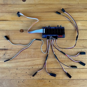 Könnte beinhalten: Eine blaue Leiterplatte mit mehreren schwarzen Steckern und Drähten sowie einem Flachbandkabel. Die Platine trägt den Text "P-CAP LOGIC" und befindet sich auf einem Holzgrund. Verschiedene schwarze Stecker sind an den Drähten befestigt.