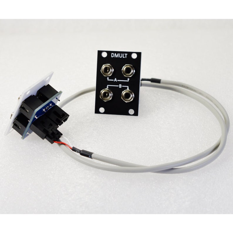 Pu&ograve; includere: Componente elettronico con un pannello nero etichettato "DMULT" con quattro jack audio. Un connettore bianco con pi&ugrave; porte nere &egrave; collegato al pannello tramite un cavo grigio. L'immagine &egrave; su sfondo bianco.