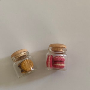 Peut inclure: Deux bocaux en verre avec des couvercles en bois, l'un rempli de biscuits roses et blancs, l'autre de biscuits bruns et jaunes.