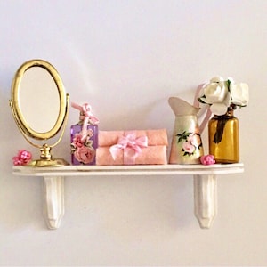 Könnte beinhalten: Ein Miniatur-Waschtischregal mit einem goldgerahmten Spiegel, einer lila Parfümflasche mit floralen Akzenten, zwei rosa Handtüchern mit Schleife, einem kleinen Krug mit Blumenmuster und einer braunen Glasvase mit weißen Blumen. Das Regal ist weiß.