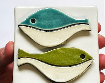 May include: A square ceramic tile featuring two stylized fish. The fish are teal and green with black eye details, set against a white background. The tile has a glossy finish, and the fish design is raised.
