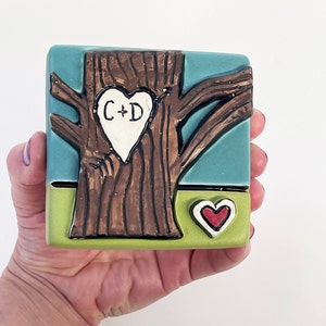 May include: A ceramic tile with a brown tree with a white heart carved into it. The heart has the initials "C+D" inside. The tile has a green base and a blue sky background. There is a small red heart at the bottom of the tile.