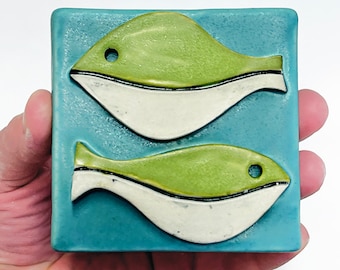 May include: A ceramic tile with two green and white fish on a blue background.