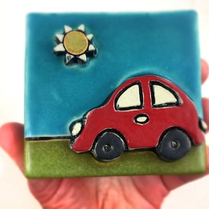 May include: A red car with a white windshield and black wheels is driving on a green field under a blue sky with a yellow sun.