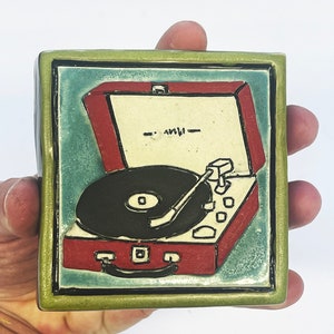 May include: A red and white ceramic tile with a green border, depicting a vintage record player with a black vinyl record. The record player has a white lid that is open, revealing the record player's interior.