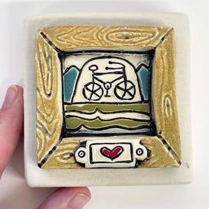 May include: A square ceramic tile with a bicycle scene framed in a wood-grain border. The artwork features a black bicycle, blue mountains, and green water. A small rectangular tag with a red heart is at the bottom.