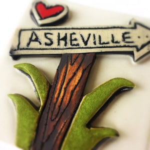 May include: A white ceramic tile with a brown wooden signpost pointing to the right with the text "ASHEVILLE" on it. A red heart is above the signpost and green leaves are below it.