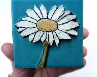 May include: A white daisy with a yellow center is painted on a teal blue ceramic tile.
