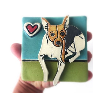May include: A ceramic tile with a hand-painted image of a white and brown dog with black markings. The dog is sitting on a green field with a blue sky behind it. A red heart is in the upper left corner of the tile.
