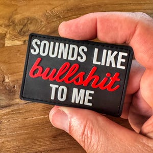 以下が含まれることがあります： 「SOUNDS LIKE bullshit TO ME」の文字が白と赤で書かれた黒い長方形のパッチ。パッチは手で持たれています。