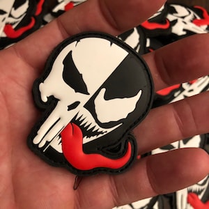 May include: A black, white, and red patch featuring a skull design with a split face. One side is a white skull, the other is a black and white design with a red tongue. The patch is likely for a jacket or bag.