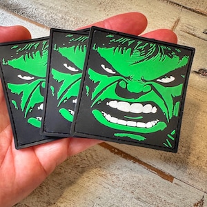 以下が含まれることがあります： 黒い背景に緑と白のハルクの顔が描かれた正方形のパッチが3つ。ハルクの怒った表情が際立っており、歯をむき出しにし、眉をひそめています。パッチは手で持たれています。