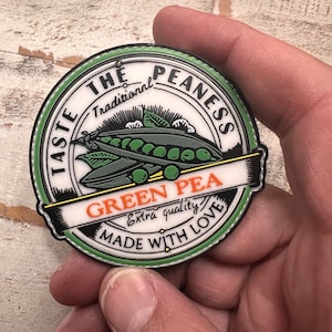 May include: A green and white round sticker with the text "Taste the Pea-ness Traditional Green Pea Extra quality Made with Love" in a circle. The sticker features a cartoon image of a pea pod with peas inside.