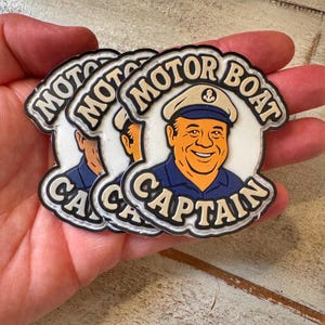 以下が含まれることがあります： モーターボートキャプテンのステッカーの束。各ステッカーには、白い帽子と濃い青色の制服を着た、笑顔の漫画の船長が描かれています。「MOTOR BOAT CAPTAIN」の文字は、黒い縁取りのあるベージュ色のフォントです。