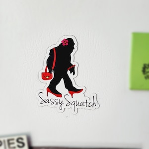 May include: A black silhouette of a sasquatch wearing red high heels, a red purse, and a pink flower in her hair. The text "Sassy Squatch" is written in black cursive below the silhouette.