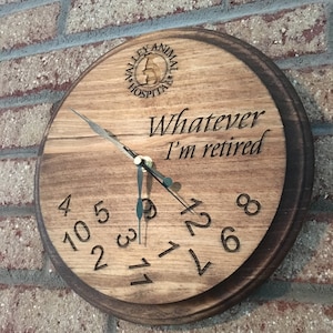 May include: A wooden clock with a dark brown stain. The clock face has a white background with black numbers and hands. The clock is decorated with the text "Whatever I'm retired".