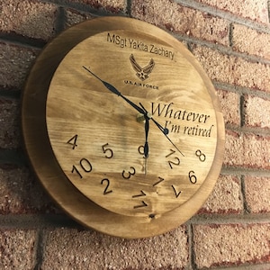 May include: A round wooden clock with a light brown finish, featuring engraved numerals and hands. The clock face includes the text "Whatever I'm retired" and the U.S. Air Force emblem. The clock is mounted on a brick wall.