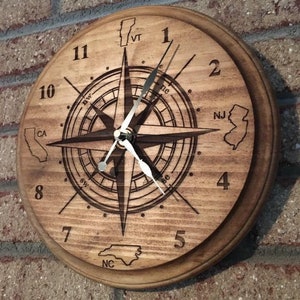Op de afbeelding: Een houten klok met een kompasontwerp en omtrekken van staten rond de cijfers. De staten zijn Vermont, Californië, New Jersey en North Carolina.