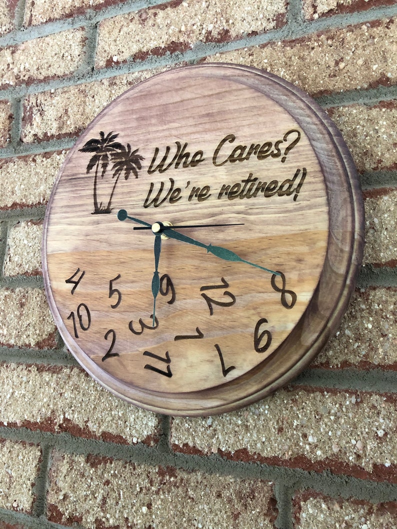 Op de afbeelding: Een houten klok met een bruine houten afwerking. De wijzerplaat van de klok heeft zwarte cijfers en wijzers. De klok heeft een palmboomontwerp en de tekst "Who Cares? We're retired!"